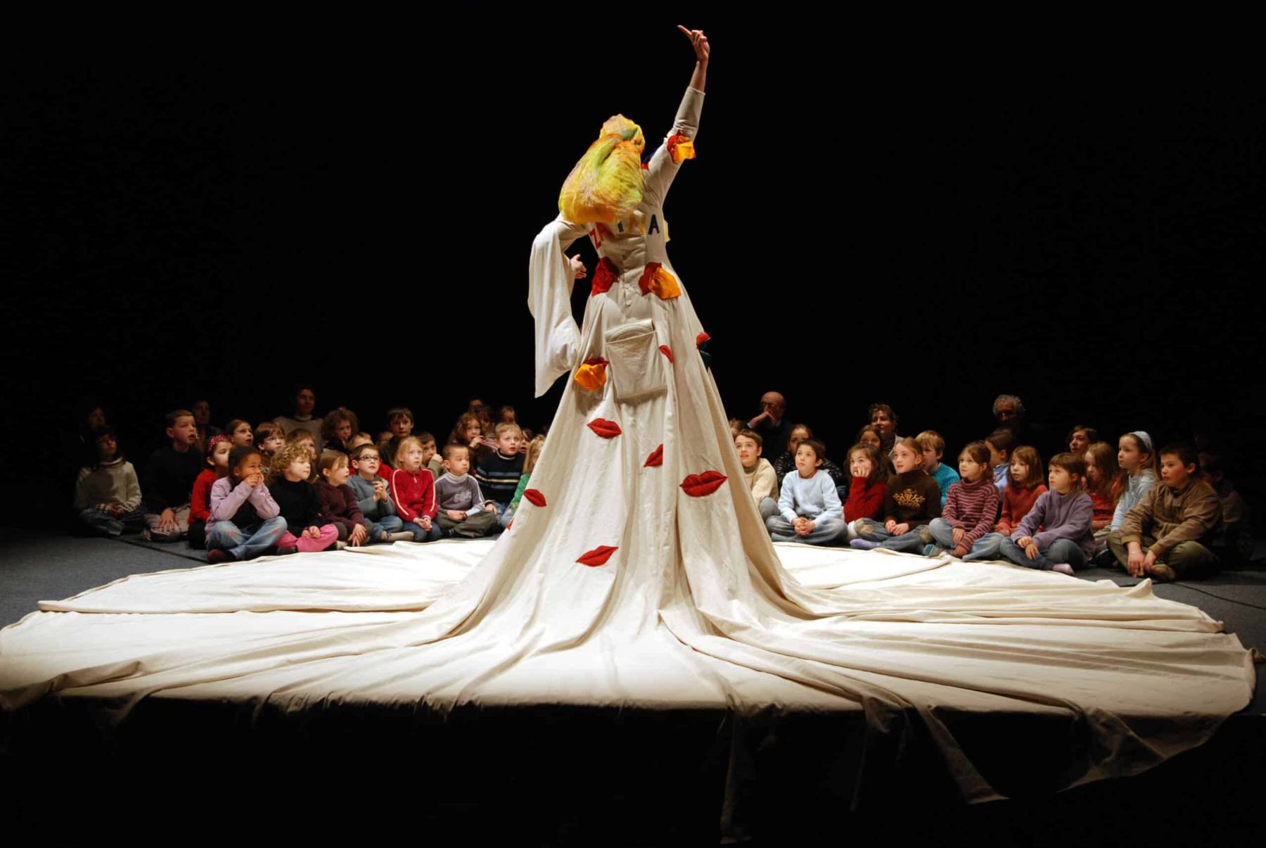 La chanteuse, habillée d'une grande robe, sur laquelle sont cousues des grandes bouches, chantent devant des enfants assis en tailleur, autour de sa grande robe